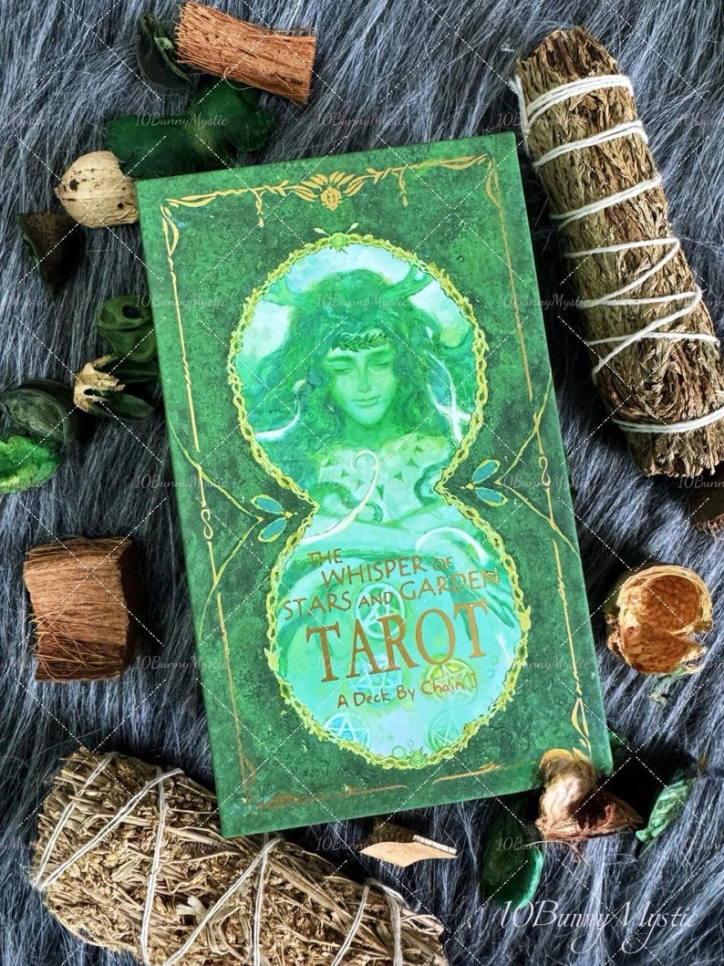 May include: A green tarot card deck titled "The Whisper of the Stars and Garden Tarot" with a watercolor illustration of a woman. The deck is surrounded by dried herbs and decorative elements on a gray fur-like surface.