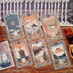 K&ouml;nnte beinhalten: Eine Sammlung von sechs rechteckigen Tarotkarten mit Illustrationen von Sternen, einem Storch, einem Hund, einem Turm, einem Garten und einem Berg. Jede Karte zeigt eine Nummer, ein Spielkartensymbol und einen Titel auf Englisch. Die Karten sind auf einer Holzoberfl&auml;che angeordnet, mit einer Teilansicht einer siebten Karte und einer aufgerollten Schriftrolle.