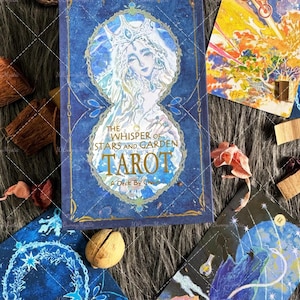 May include: A blue tarot card deck titled "The Whisper of Stars and Garden Tarot" with a celestial illustration of a woman. Several tarot cards with various illustrations are scattered around the deck, along with dried flowers and a nut.