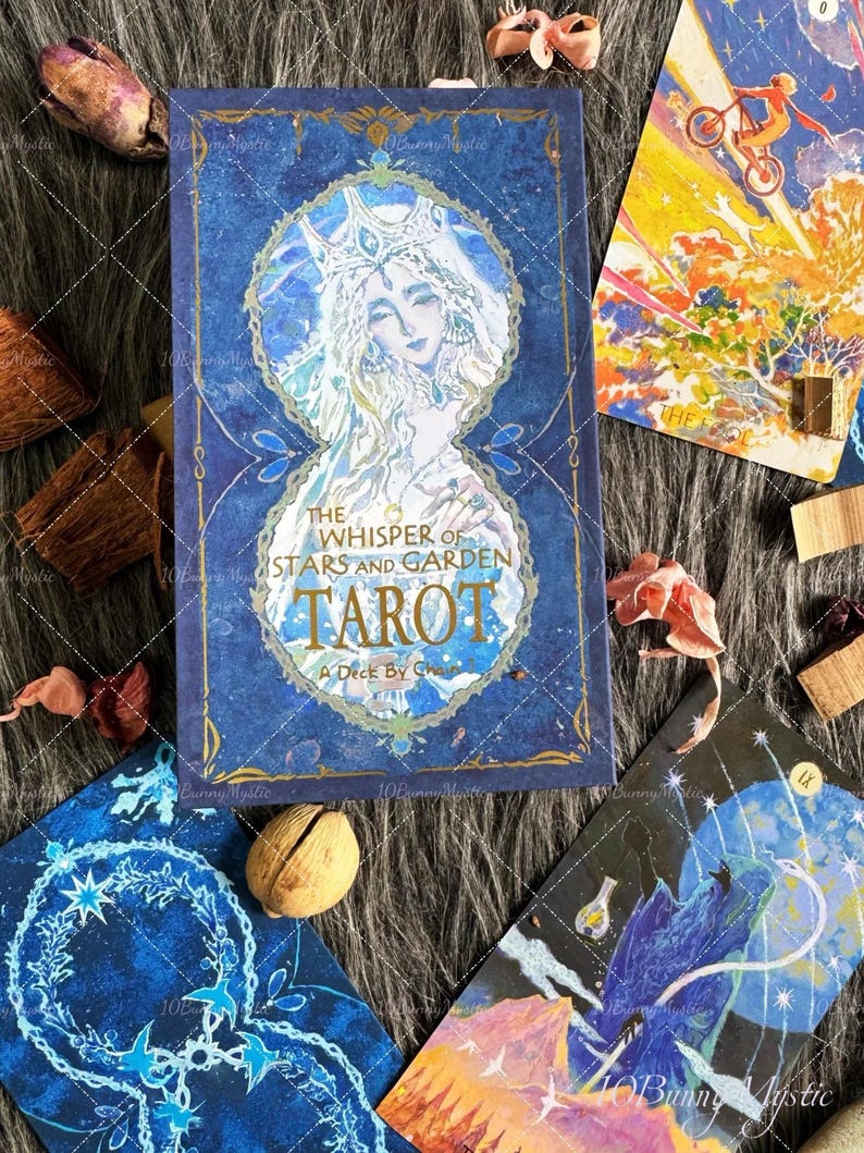 May include: A blue tarot card deck titled "The Whisper of Stars and Garden Tarot" with a celestial illustration of a woman. Several tarot cards with various illustrations are scattered around the deck, along with dried flowers and a nut.