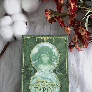 May include: A tarot card deck titled "The Whisper of Stars and Garden Tarot" with a green and gold design. The box features an illustration of a woman with long hair. The deck is surrounded by cotton and flowers.