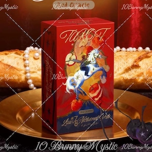 May include: A red tarot card box with gold lettering and a colorful illustration of a horse and rider. The box is on a gold plate with bread, pearls, and cutlery in the background. The text "Tarot" is visible.