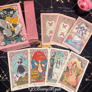 May include: A pink Transformation Tarot card deck with a matching box. The box features a fairy illustration and the words "Transformation Tarot." Several cards are displayed, each with unique illustrations and titles like "The Fool" and "The Empress."