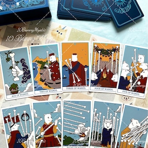 May include: A deck of tarot cards featuring illustrations of white bunnies in various scenes, labelled from Ace of Wands to Ten of Wands. The cards have blue borders and are displayed alongside a matching blue box with a bunny illustration on the lid.