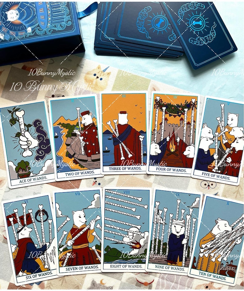 May include: A deck of tarot cards featuring illustrations of white bunnies in various scenes, labelled from Ace of Wands to Ten of Wands. The cards have blue borders and are displayed alongside a matching blue box with a bunny illustration on the lid.