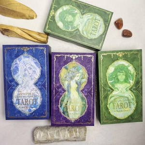 May include: Tarot card decks in blue, purple, and green boxes with gold accents. Each box features a watercolor illustration of a woman and the text "The Whisper of Stars and Garden Tarot". Additional items include a feather, a stick, and small stones.