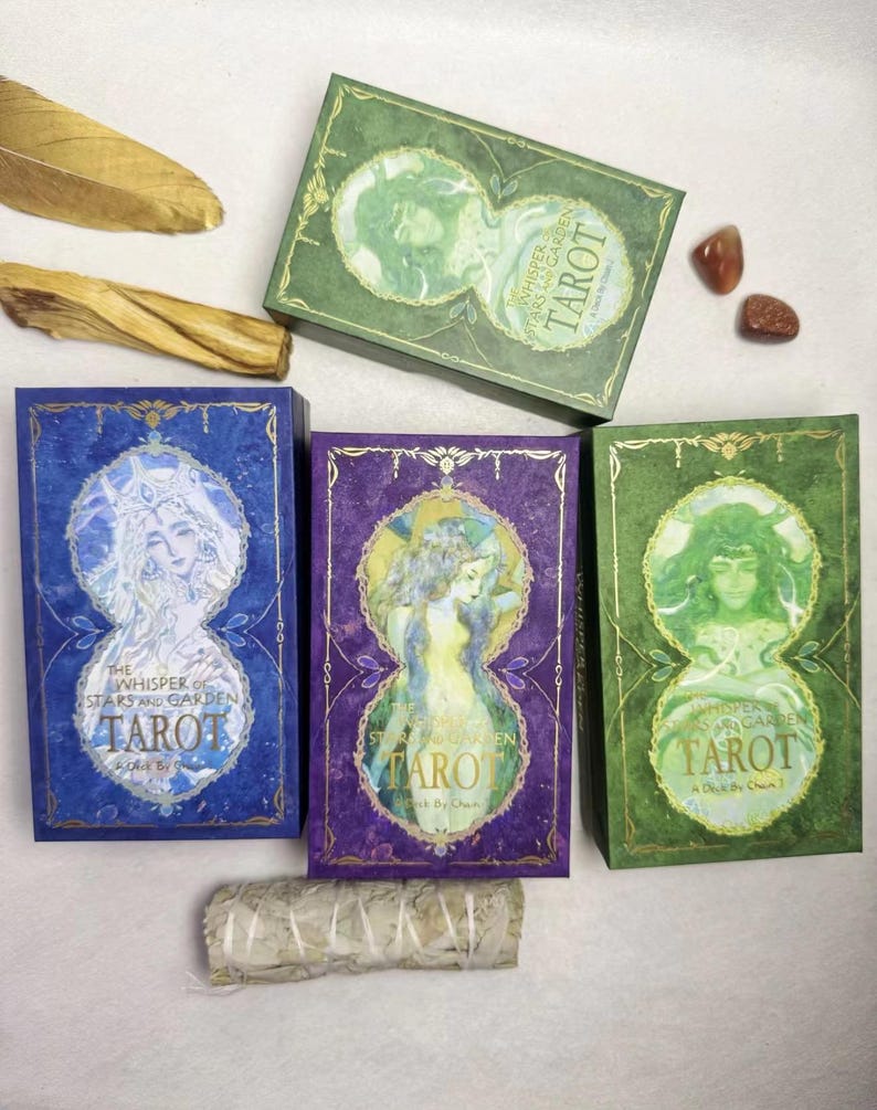 May include: Tarot card decks in blue, purple, and green boxes with gold accents. Each box features a watercolor illustration of a woman and the text "The Whisper of Stars and Garden Tarot". Additional items include a feather, a stick, and small stones.