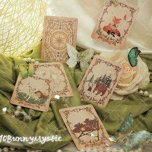 May include: A selection of vintage tarot cards featuring illustrations of a fox, bear, and Dalmatian, set against a backdrop of green fabric. The cards have decorative borders and are accompanied by butterflies and a rose.