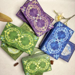 May include: A collection of tarot cards in shades of green, blue, and purple. Each card features a stylized infinity symbol with floral and star designs. The cards are arranged in stacks and fanned out, showcasing the intricate artwork.