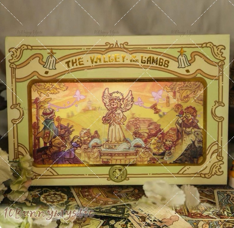 May include: A decorative box with the text "THE VALLEY OF THE LAMBS" features an illustration of an angel and figures in a landscape. The box has a gold and green border with ornate details and a small emblem at the bottom.