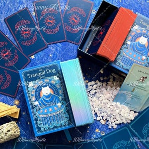 May include: A tarot deck titled The Tranquil Dog featuring a blue box with a white dog illustration, a deck with iridescent edges, and a stack with orange edges. Several cards show a red bone symbol, and a small card displays the text Like it, Just.