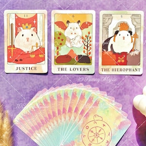 May include: Three tarot cards featuring guinea pig illustrations labeled Justice, The Lovers, and The Hierophant, displayed above a fanned deck of cards with a pastel watercolor background and a circular symbol on the back of the cards.