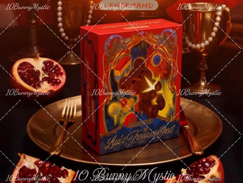 May include: A red gift box with a teddy bear illustration and the text "Luv's Treasure Chest" sits on a gold plate with a fork and knife. A halved pomegranate and a gold goblet are in the background.