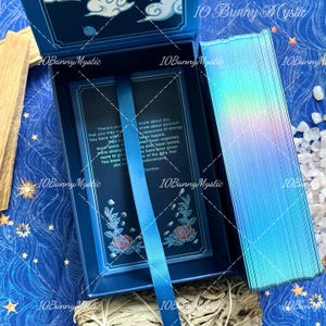 May include: A rectangular blue box with a hinged lid featuring white cloud illustrations and the text 10 Bunny Mystic. Inside, a blue ribbon sits over a card with printed text and rose graphics. A deck of cards with iridescent blue edges is placed beside the box.