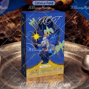 May include: A blue tarot card box with the word "TAROT" in white script. The box features a celestial illustration with a figure, stars, and butterflies. The text "Cosmic Fold" is visible. A fork and spoon flank the box on a silver platter.