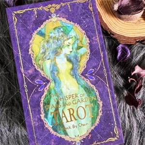 May include: A rectangular tarot deck box with a deep purple background and gold filigree borders. The centre features an oval frame containing an illustration of a figure with flowing hair. Text reads: The Whisper of Stars and Garden Tarot, A Deck By Chain.