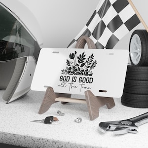 Puede incluir: Placa de matrícula blanca con el texto "GOD IS GOOD all the Time" y un diseño floral. La placa se muestra sobre un soporte de madera, con una bandera de carreras y neumáticos en el fondo. También se ven un casco, herramientas y llaves.