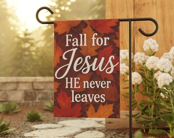 May include: A garden flag with the text "Fall for Jesus HE never leaves" in white script. The flag features a background of autumn leaves in shades of red, orange, and brown. The flag is displayed on a black metal stand.