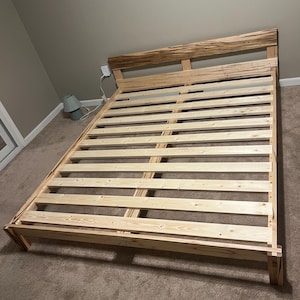 May include: A wooden bed frame with a slatted base and a headboard. The headboard features a distinctive, textured pattern. The frame is constructed from light-coloured wood and is positioned on a carpeted floor. The bed frame is designed to accommodate a mattress.