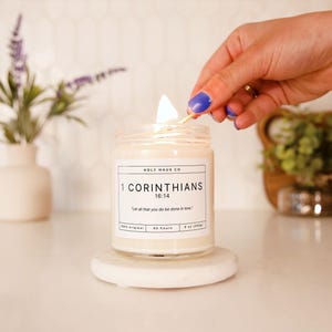 May include: A lit candle in a clear glass jar on a white marble coaster. The candle has a label with the text "1 CORINTHIANS 16:14" and "Let all that you do be done in love." The candle is 9 oz (255g) and has a 60-hour burn time.