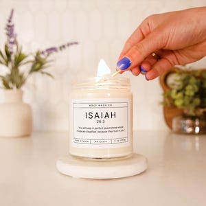 Puede incluir: Una vela encendida en un tarro de vidrio transparente sobre un posavasos de mármol blanco. La vela tiene una etiqueta blanca con el texto "ISAIAH 26:3". La vela es de 255g y dura 60 horas. Una mano con esmalte de uñas azul enciende la vela con una cerilla.