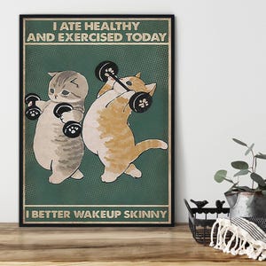 Könnte beinhalten: Ein gerahmtes Poster mit grünem Hintergrund zeigt zwei Cartoon-Katzen beim Gewichtheben. Der Text oben lautet "I ATE HEALTHY AND EXERCISED TODAY" und der Text unten lautet "I BETTER WAKEUP SKINNY".