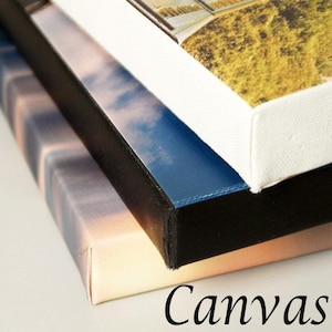 May include: Close-up of three stretched canvas prints stacked on top of each other. The canvases feature images of a sunset, a blue sky with clouds, and a landscape. The edges are black, white, and blue. The word "Canvas" is written in black.