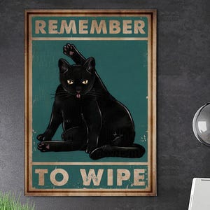 Puede incluir: Un póster enmarcado con una ilustración de un gato negro. El gato se está acicalando, con la lengua fuera. El póster tiene las palabras "REMEMBER TO WIPE" en una fuente vintage, sobre un fondo turquesa.