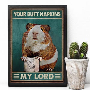 May include: Framed poster featuring a guinea pig holding a roll of toilet paper. The poster has the text "YOUR BUTT NAPKINS" above the guinea pig and "MY LORD" below. The background is teal with a distressed texture.