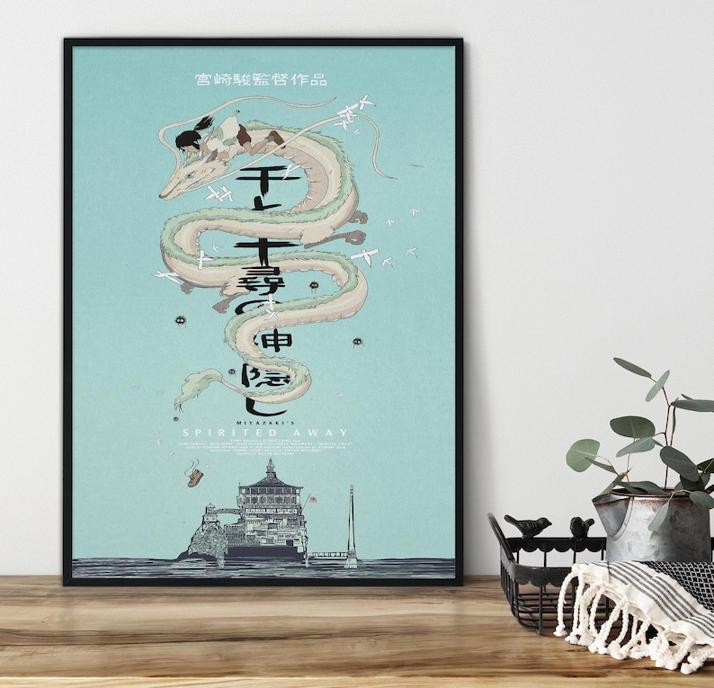 May include: Framed poster with artwork from the film "Spirited Away." It features a serpentine dragon, Japanese text, and a building on an island, set against a light blue backdrop. The poster is in a black frame.
