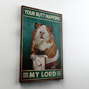 May include: A framed poster with the text "YOUR BUTT NAPKINS" at the top and "EMY LORDE" at the bottom. The poster features a guinea pig holding a roll of toilet paper. The guinea pig has brown and white fur.