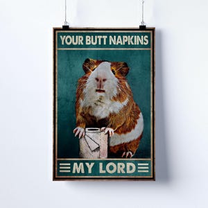 May include: A framed poster featuring a guinea pig with brown and white fur, holding a roll of toilet paper. The poster has the text "YOUR BUTT NAPKINS" at the top and "EMY LORD" at the bottom, set against a teal background.