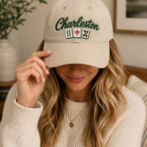 Peut inclure: Une casquette de baseball beige avec le mot Charleston brodé en écriture cursive vert foncé. Sous le texte se trouvent trois tuiles de mah-jong, dont deux tuiles bambou vertes et une tuile caractère rouge, brodées en rangée horizontale.