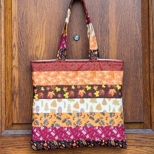 May include: A quilted tote bag with a patchwork design featuring autumn colors and leaf patterns. The bag has a rectangular shape with a wide base and two handles. The color palette includes shades of orange, brown, and burgundy.