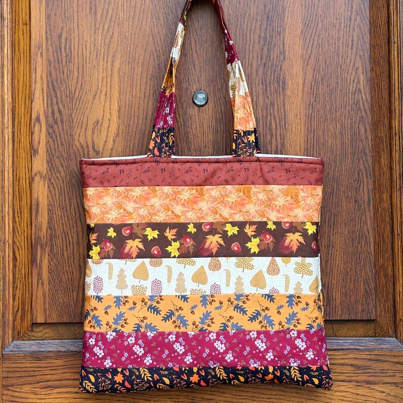 May include: A quilted tote bag with a patchwork design featuring autumn colors and leaf patterns. The bag has a rectangular shape with a wide base and two handles. The color palette includes shades of orange, brown, and burgundy.