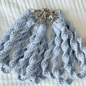 May include: Several light blue macrame keychains with silver-colored clasps. The keychains are made of twisted, braided cotton cord. The keychains are arranged in a fan shape, with the clasps clustered together.