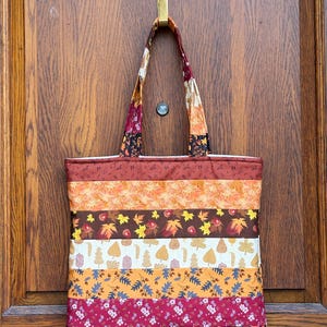 May include: A colorful tote bag with a patchwork design, featuring horizontal stripes of various autumn-themed fabrics. The bag includes patterns of leaves, flowers, and berries in shades of orange, brown, and burgundy. The handles are made of a matching fabric.
