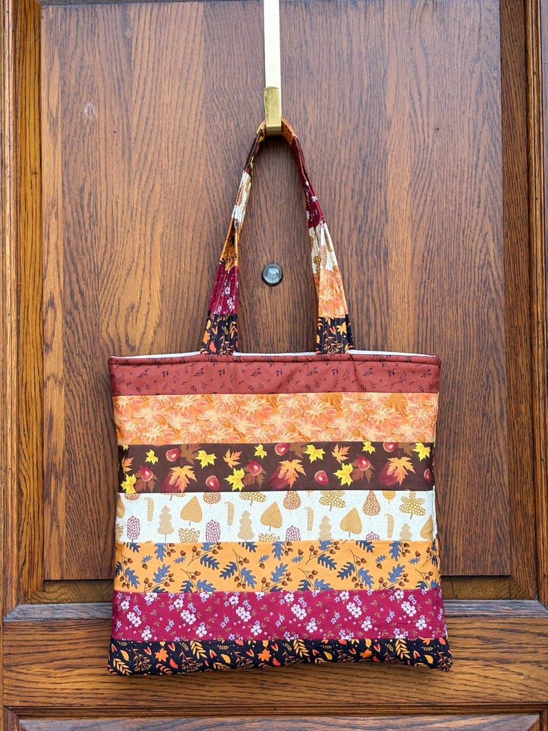 May include: A colorful tote bag with a patchwork design, featuring horizontal stripes of various autumn-themed fabrics. The bag includes patterns of leaves, flowers, and berries in shades of orange, brown, and burgundy. The handles are made of a matching fabric.