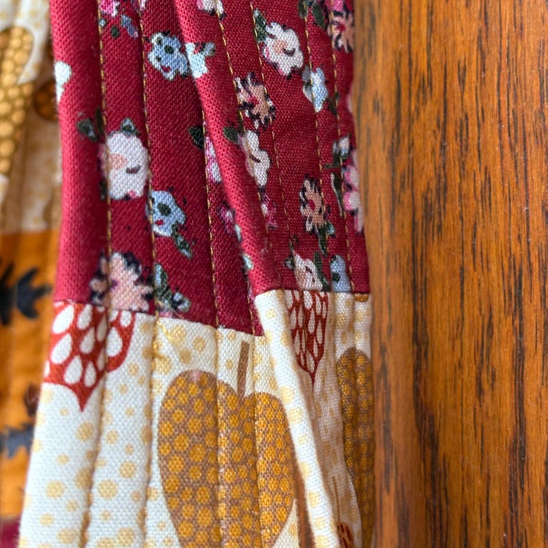 May include: Close-up of a quilted fabric with a floral pattern in red, white, and blue. The fabric also features a pattern of yellow hearts and a cream-colored background. The stitching is visible, adding texture to the design.