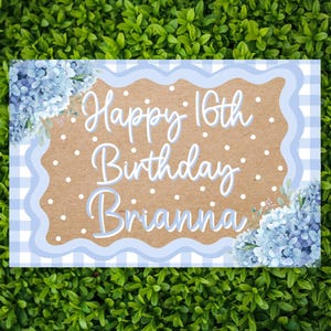 May include: A rectangular sign featuring a light blue and white gingham border with a scalloped edge. The center is tan with white polka dots and the text Happy 16th Birthday Brianna in white script. Blue hydrangea flowers decorate two corners.