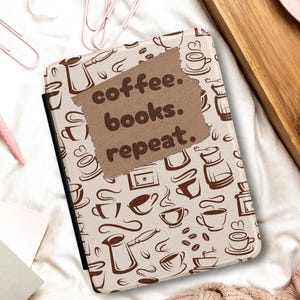 以下が含まれることがあります： 「coffee. books. repeat.」の文字とコーヒーをテーマにした模様が入ったタブレットケースが白い表面に置かれ、開いた本が隣にあります。コーヒーカップ、クロワッサン、ピンクのペン、ピンクの毛布、バラも見えます。