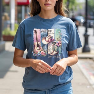 May include: A blue t-shirt featuring a Nashville-themed graphic. The design includes a guitar, cowboy hat, boots, and musical notes, with the text "Nashville" and "Girls Tour 2026". The shirt is a comfortable, casual style.