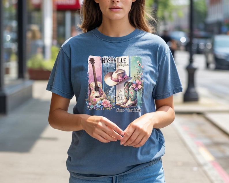 May include: A blue t-shirt featuring a Nashville-themed graphic. The design includes a guitar, cowboy hat, boots, and musical notes, with the text "Nashville" and "Girls Tour 2026". The shirt is a comfortable, casual style.