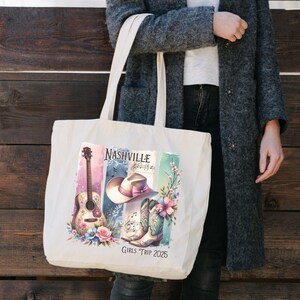 May include: Cream-colored tote bag featuring a Nashville-themed design. The design includes a guitar, cowboy hat, boots, and floral accents. The text "Nashville" and "Girls Trip 2025" are also visible. The bag is held by a person wearing a gray coat.