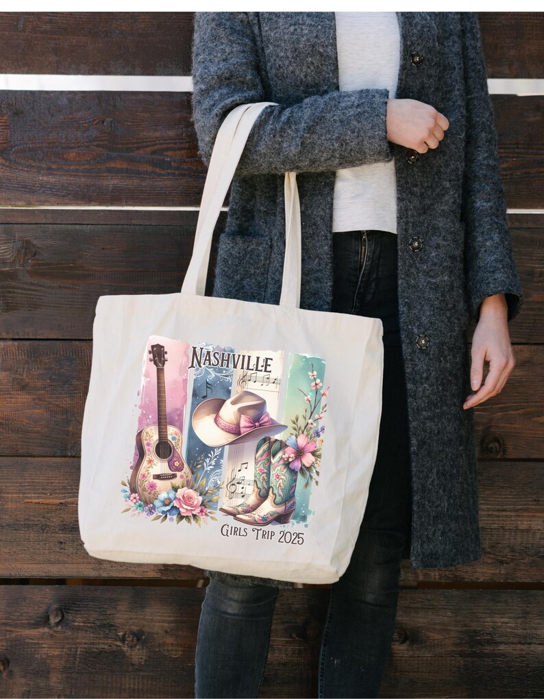May include: Cream-colored tote bag featuring a Nashville-themed design. The design includes a guitar, cowboy hat, boots, and floral accents. The text "Nashville" and "Girls Trip 2025" are also visible. The bag is held by a person wearing a gray coat.