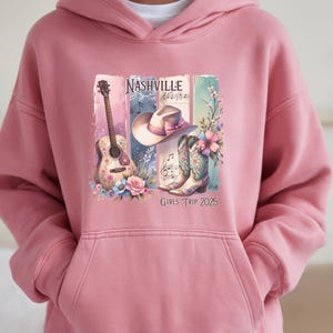 May include: A pink hooded sweatshirt featuring a graphic design with a guitar, cowboy hat, and boots. The design includes the word "Nashville" and the text "Girls Trip 2025". The graphic is in shades of pink, blue, and green.
