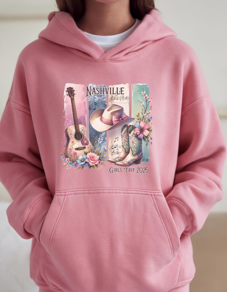 May include: A pink hooded sweatshirt featuring a graphic design with a guitar, cowboy hat, and boots. The design includes the word "Nashville" and the text "Girls Trip 2025". The graphic is in shades of pink, blue, and green.