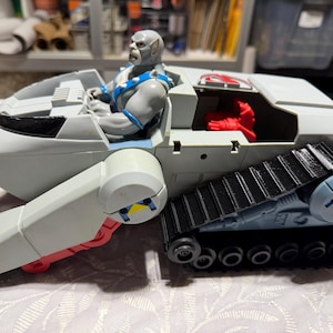 Peut inclure: Un véhicule jouet vintage, gris et noir. Il comprend une figurine grise assise avec des détails bleus et un intérieur rouge. Le véhicule a un système de chenilles de type char et provient probablement d'une gamme de jouets de science-fiction ou de figurines d'action.