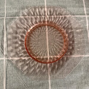 May include: A clear, octagonal glass plate with a textured surface and a pink rim. The plate has a decorative pattern radiating from the center. The plate is likely vintage or antique, and is suitable for serving food.