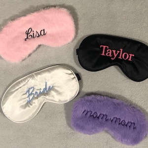 May include: Four sleep masks in the image. A pink, fluffy sleep mask with "Lisa" embroidered in black. A black satin sleep mask with "Taylor" in pink. A white satin sleep mask with "Bride" in blue. A purple, fluffy sleep mask with "mom mom" in black.